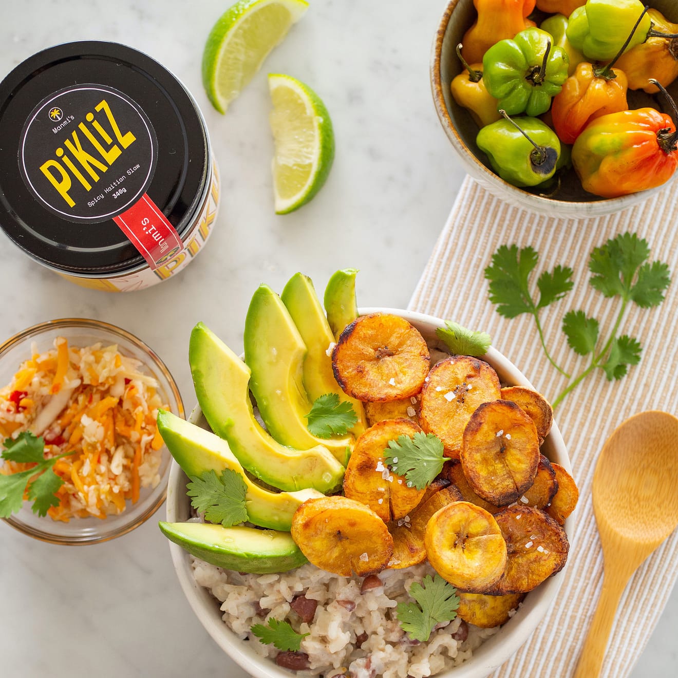 Avocado slices and fried plantains atop a bowl of rice and beans. A jar of Pikliz is also seen on the table